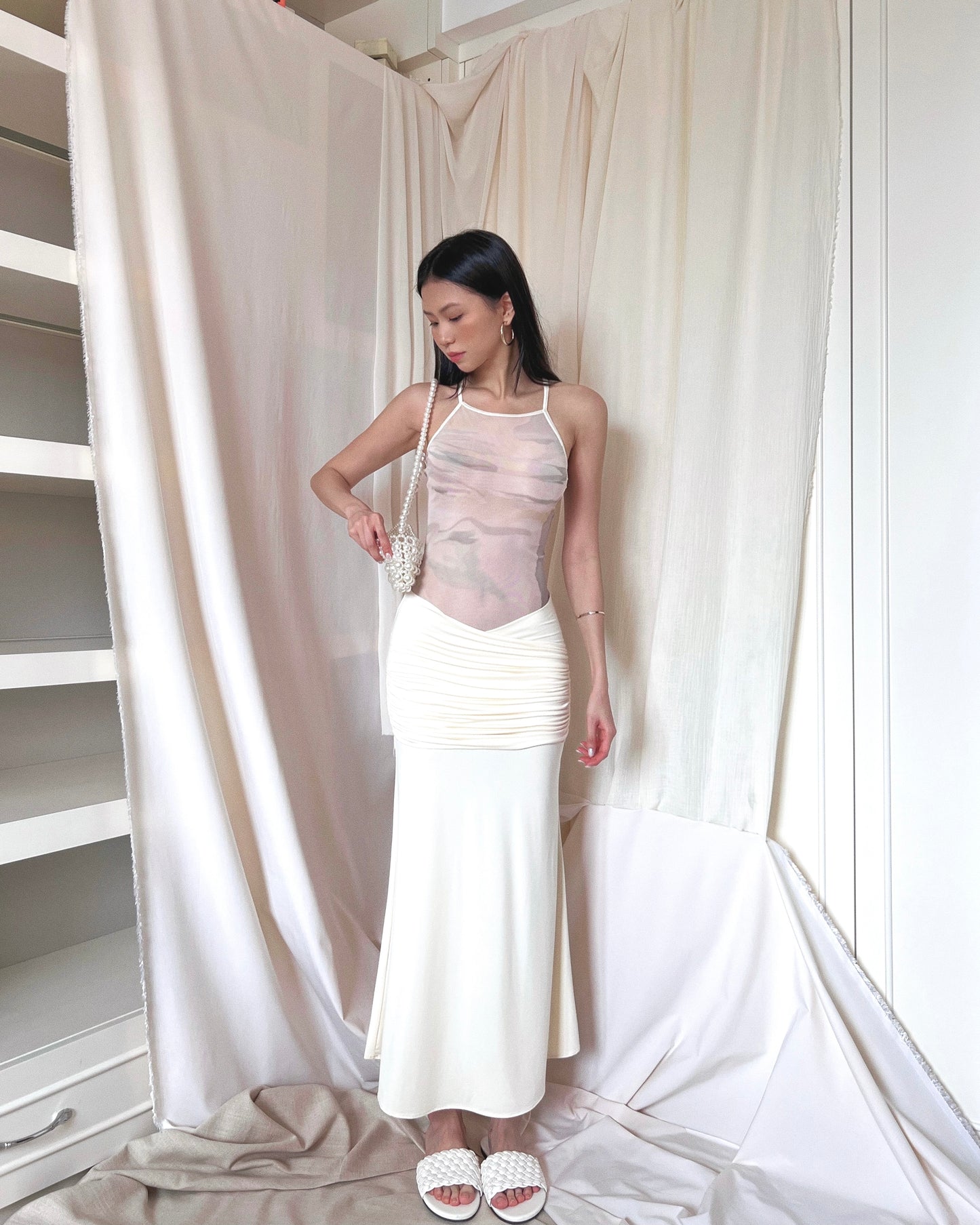 Ivory sleeveless maxi dress with printed mesh upper body panel with thin straps crossed at back and mermaid-cut draped lower part. Styled with pearl purse and white slides.