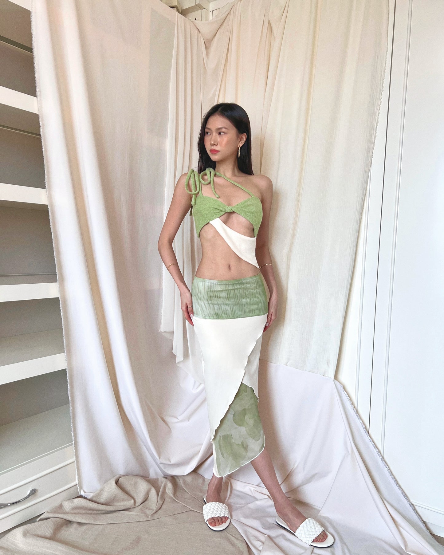 A matcha green asymmetrical knit top with a cut out style, shoulder straps to tie in different ways, and freshwater pearls around the lower back. Styled with an asymmetrical matcha green modal midi skirt with printed sheer bottom panel, printed sheer wide waistband, and white slides.
