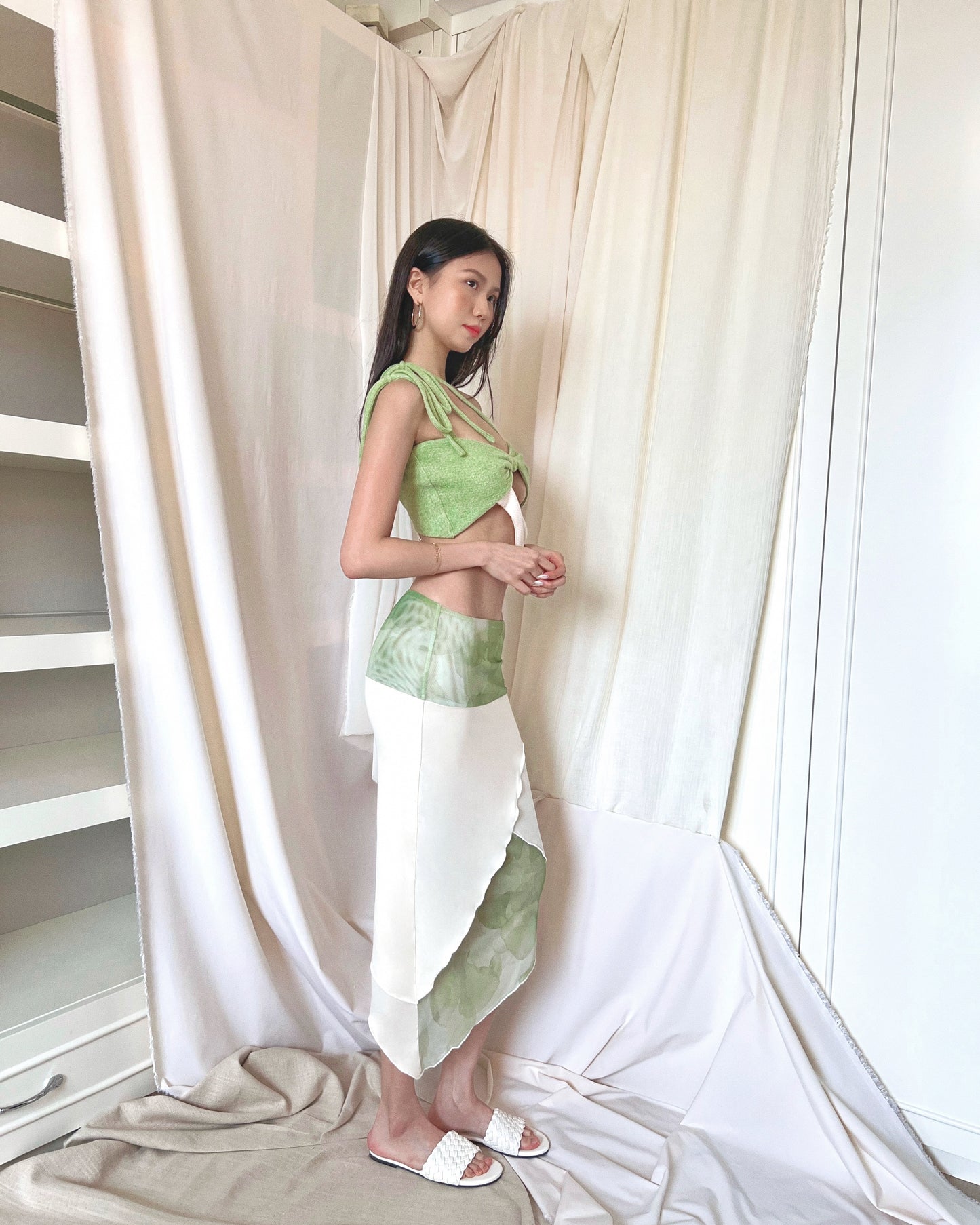 An asymmetrical matcha green modal midi skirt with printed sheer bottom panel and printed sheer wide waistband. Styled with a matcha green asymmetrical knit top with a cut out style, shoulder straps to tie in different ways, and freshwater pearls around the lower back and white slides.