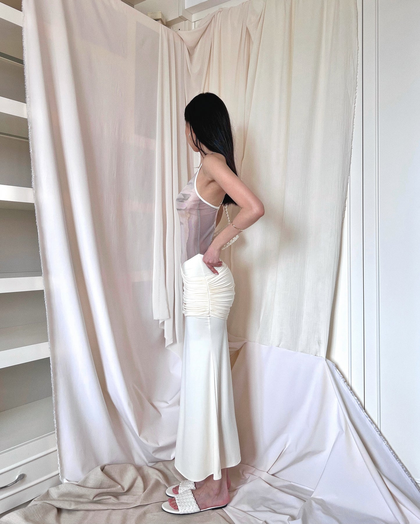 Ivory sleeveless maxi dress with printed mesh upper body panel with thin straps crossed at back and mermaid-cut draped lower part. Styled with pearl purse and white slides.