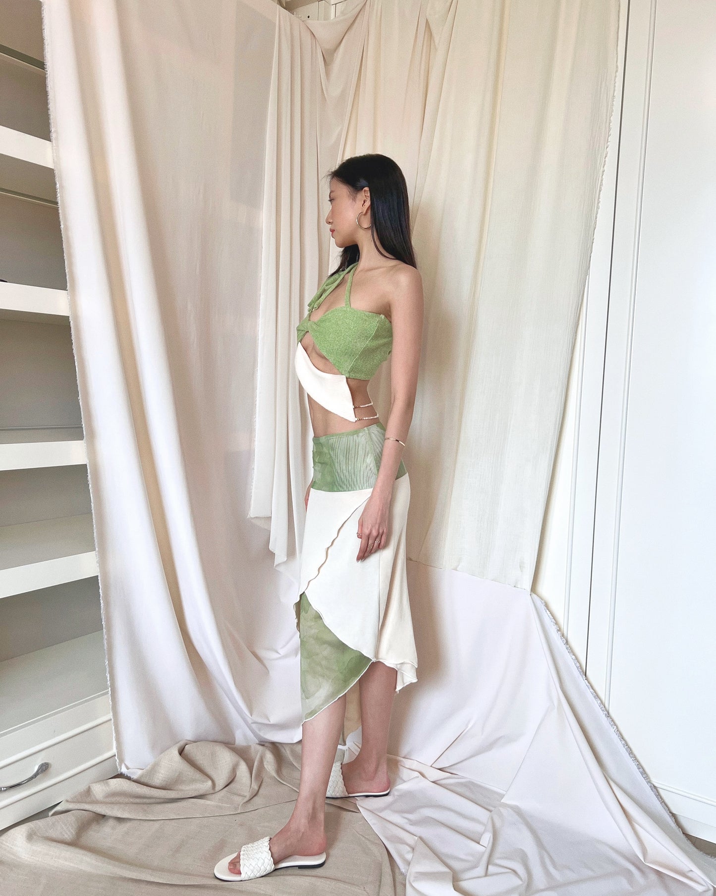 A matcha green asymmetrical knit top with a cut out style, shoulder straps to tie in different ways, and freshwater pearls around the lower back. Styled with an asymmetrical matcha green modal midi skirt with printed sheer bottom panel, printed sheer wide waistband, and white slides.