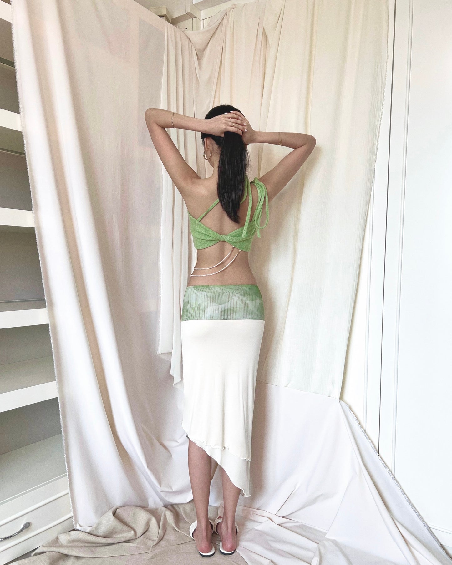A matcha green asymmetrical knit top with a cut out style, shoulder straps to tie in different ways, and freshwater pearls around the lower back. Styled with an asymmetrical matcha green modal midi skirt with printed sheer bottom panel, printed sheer wide waistband, and white slides.