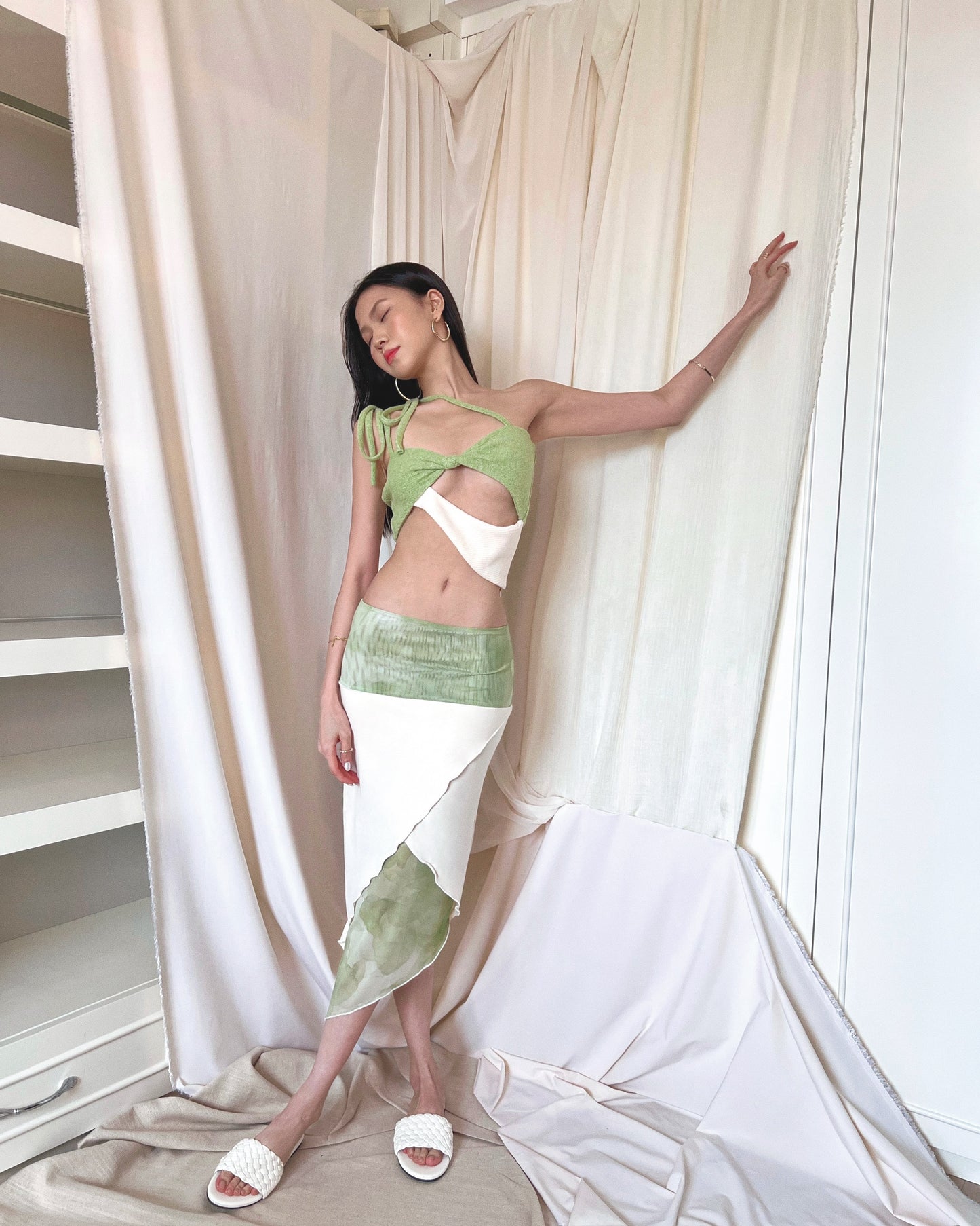 An asymmetrical matcha green modal midi skirt with printed sheer bottom panel and printed sheer wide waistband. Styled with a matcha green asymmetrical knit top with a cut out style, shoulder straps to tie in different ways, and freshwater pearls around the lower back and white slides.