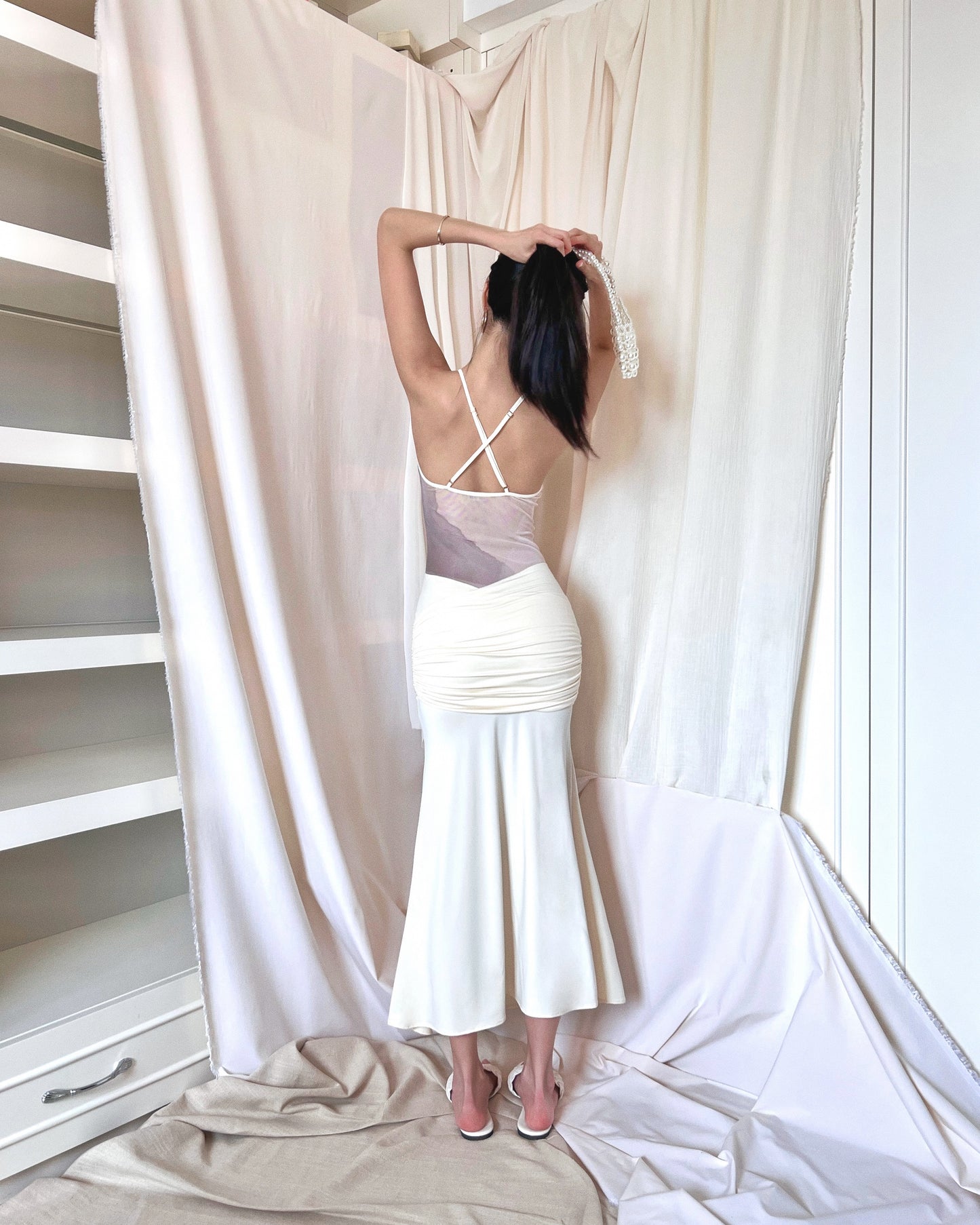 Ivory sleeveless maxi dress with printed mesh upper body panel with thin straps crossed at back and mermaid-cut draped lower part. Styled with pearl purse and white slides.