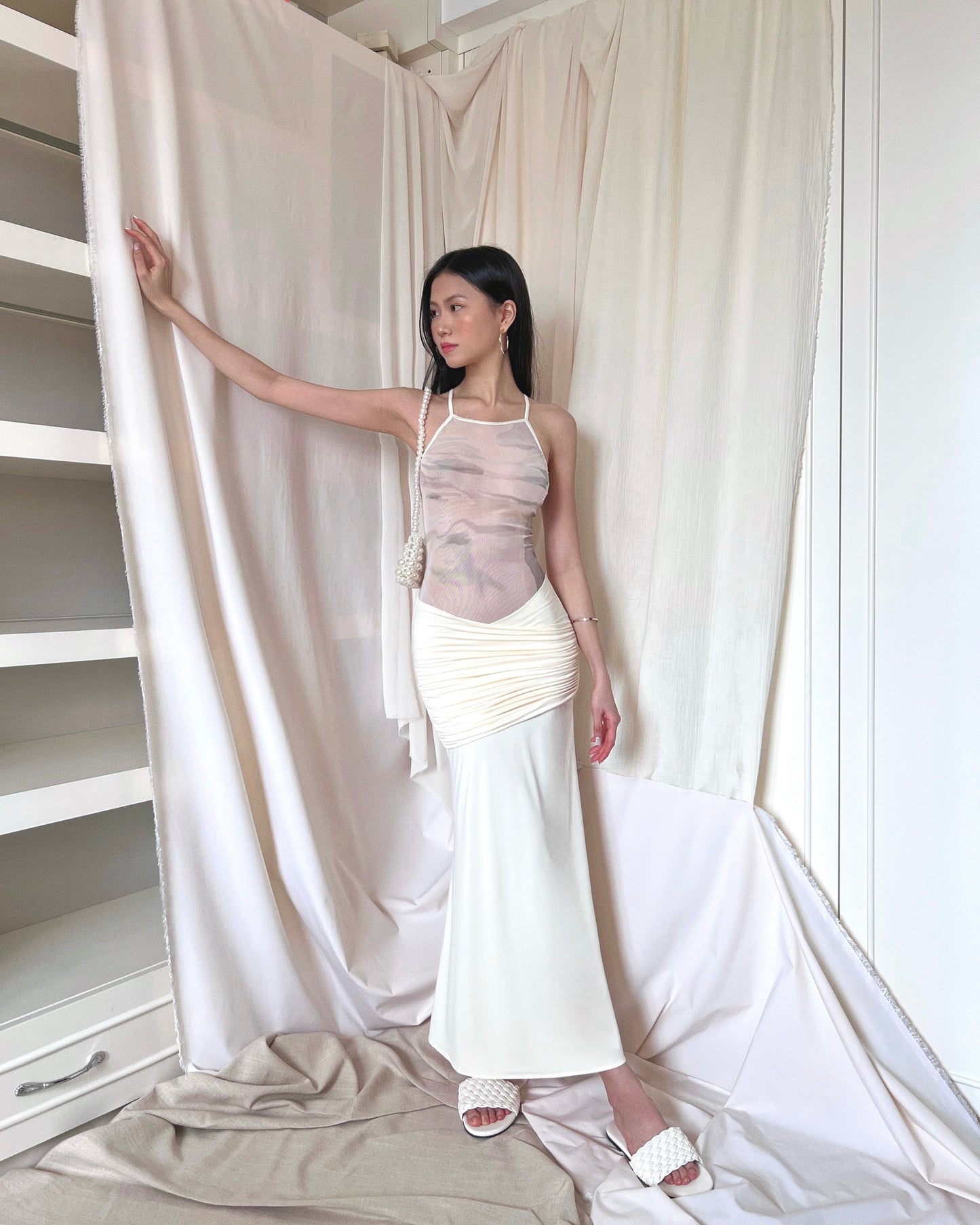 Ivory sleeveless maxi dress with printed mesh upper body panel with thin straps crossed at back and mermaid-cut draped lower part. Styled with pearl purse and white slides.