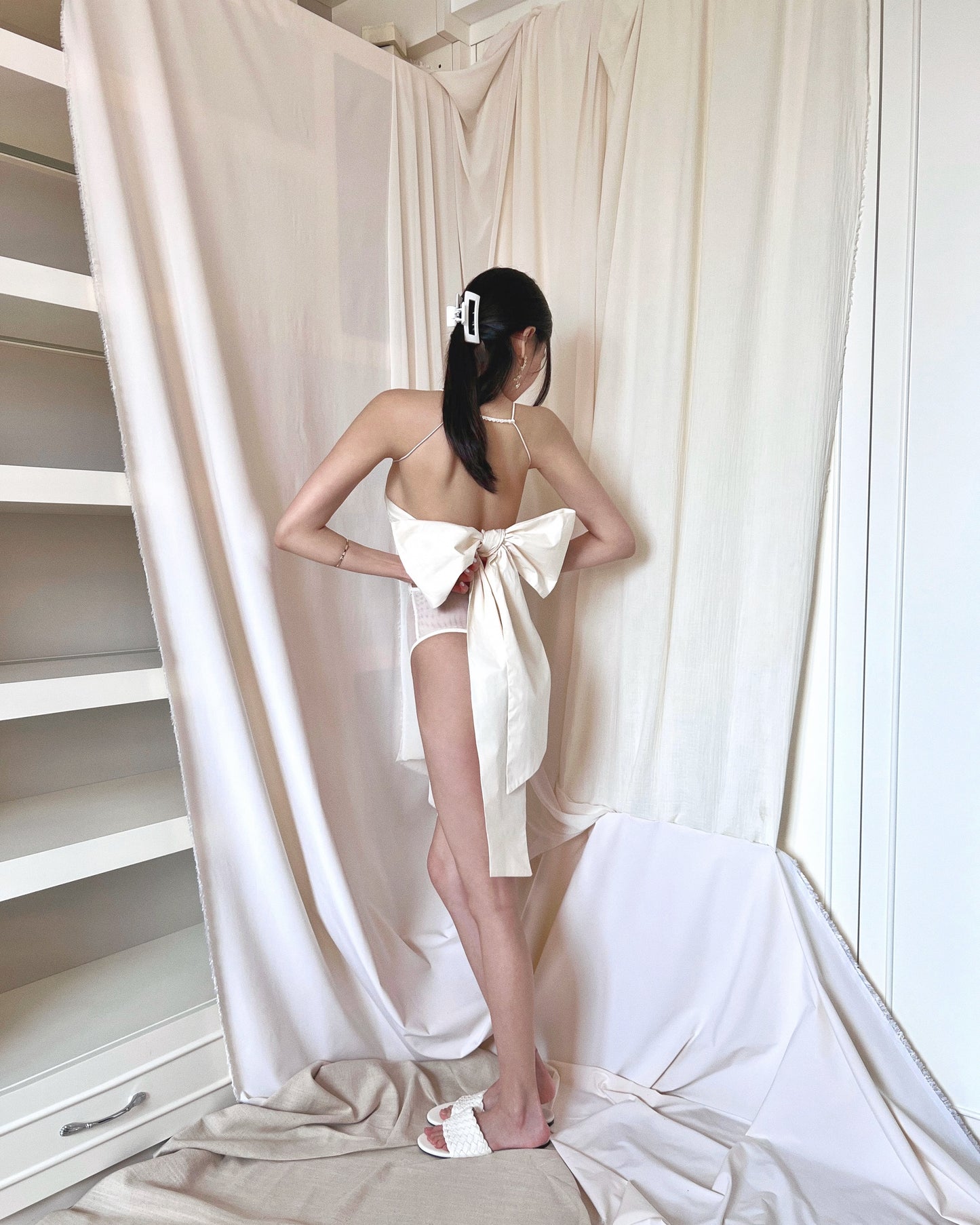 Formfitting ivory bodysuit, made in smooth and soft nylon fabric, with side mesh panels and wide and long woven back straps as bow. Styled with Jacquemus mini purse and white slides.