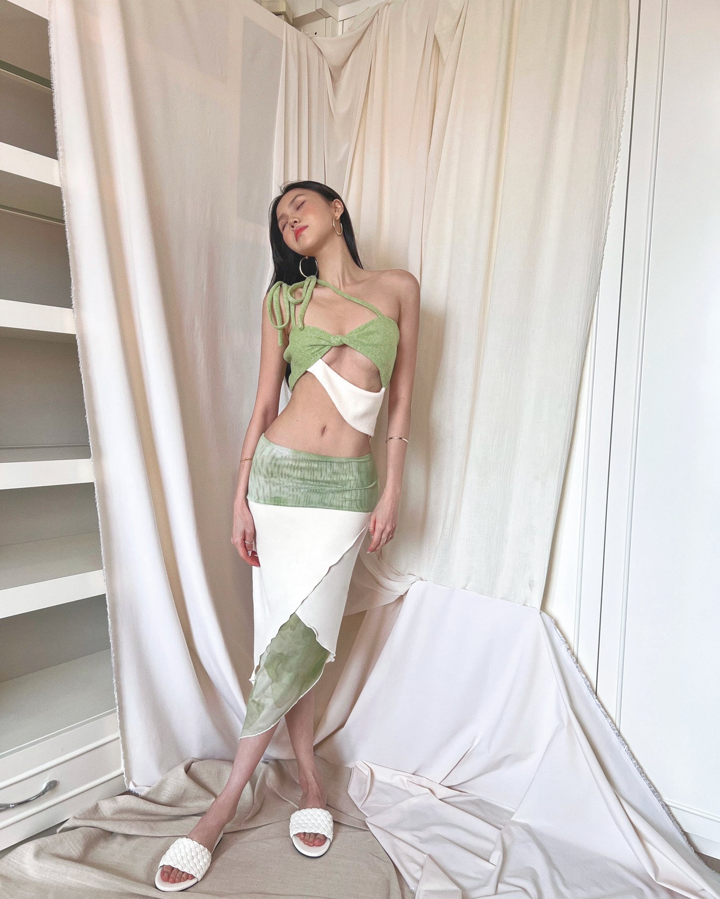 A matcha green asymmetrical knit top with a cut out style, shoulder straps to tie in different ways, and freshwater pearls around the lower back. Styled with an asymmetrical matcha green modal midi skirt with printed sheer bottom panel, printed sheer wide waistband, and white slides.