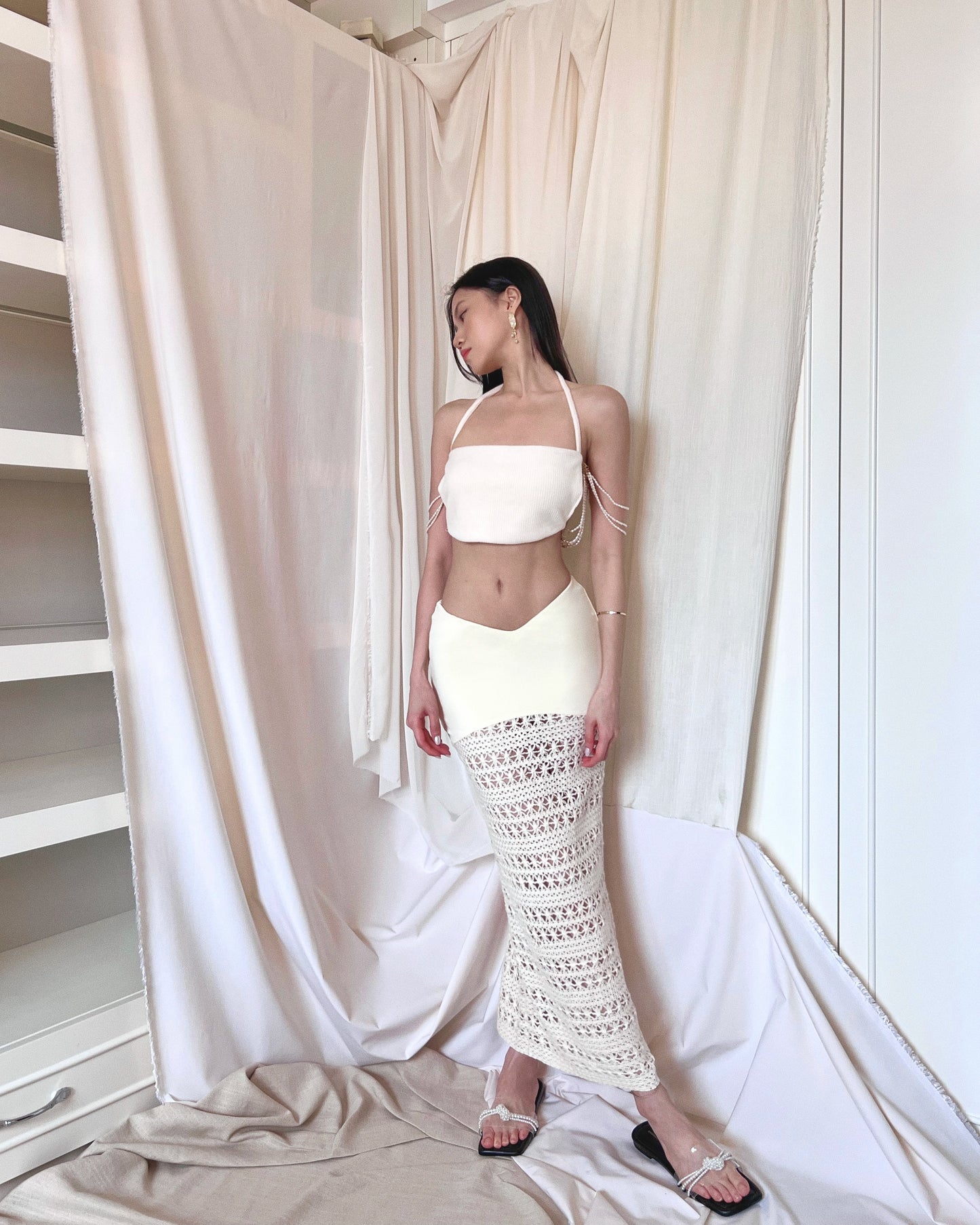 A freshwater pearl embellished sleeveless white knit top with back thin woven straps. Styled with an ivory crochet maxi skirt with nylon fabric and crochet fabric and a symmetrically diagonal waistline and pearl slides.