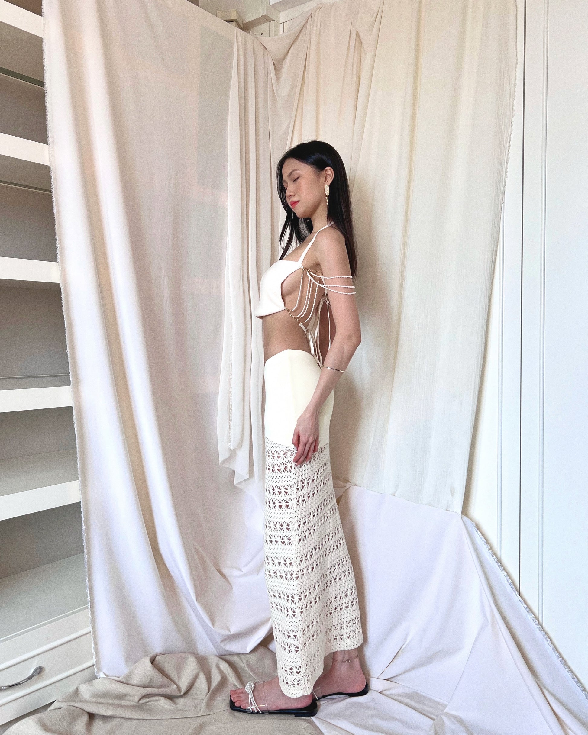 A freshwater pearl embellished sleeveless white knit top with back thin woven straps. Styled with an ivory crochet maxi skirt with nylon fabric and crochet fabric and a symmetrically diagonal waistline and pearl slides.