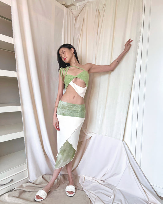 A matcha green asymmetrical knit top with a cut out style, shoulder straps to tie in different ways, and freshwater pearls around the lower back. Styled with an asymmetrical matcha green modal midi skirt with printed sheer bottom panel, printed sheer wide waistband, and white slides.
