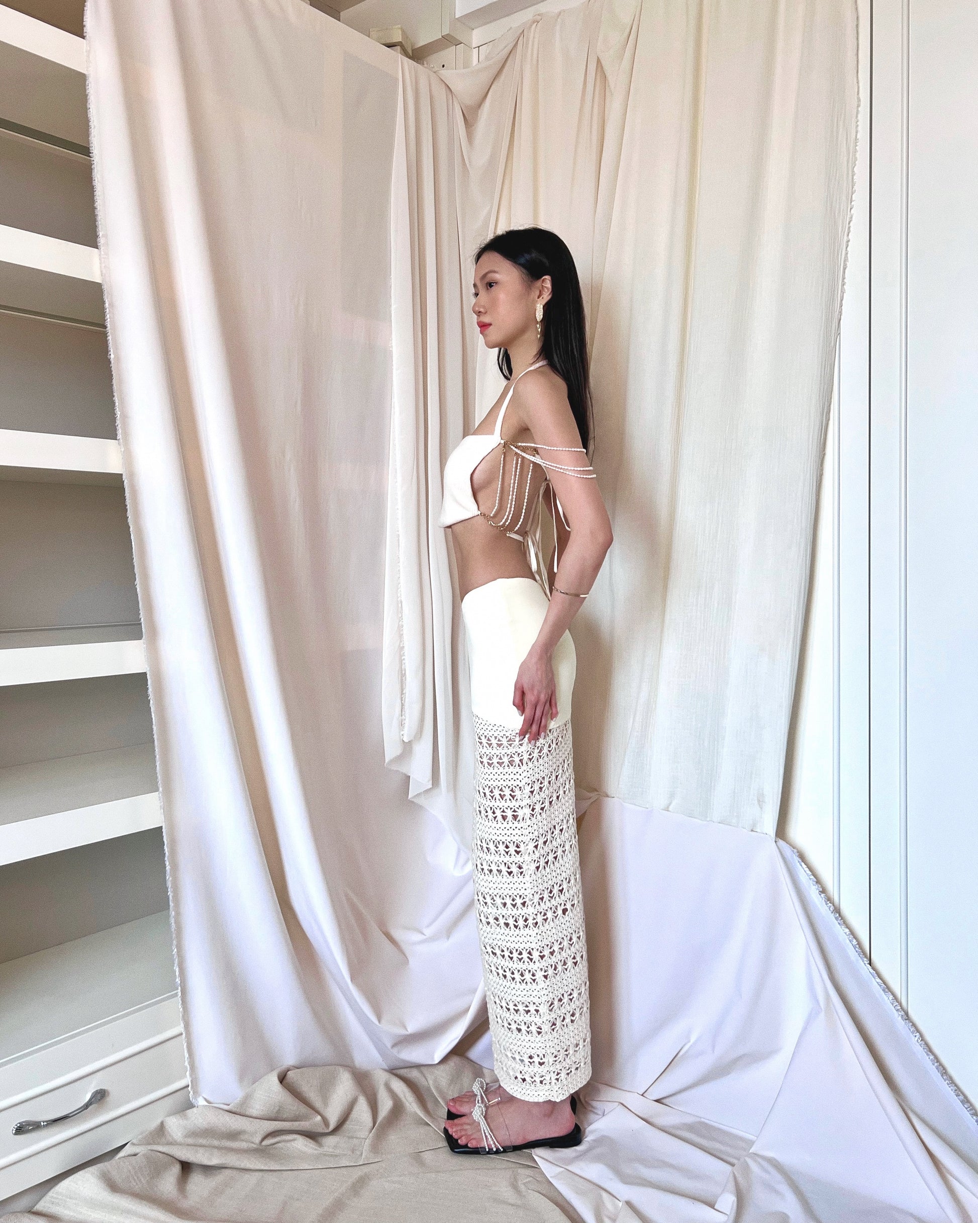 An ivory crochet maxi skirt with nylon fabric and crochet fabric and a symmetrically diagonal waistline. Styled with freshwater pearl embellished sleeveless white knit top and pearl slides.