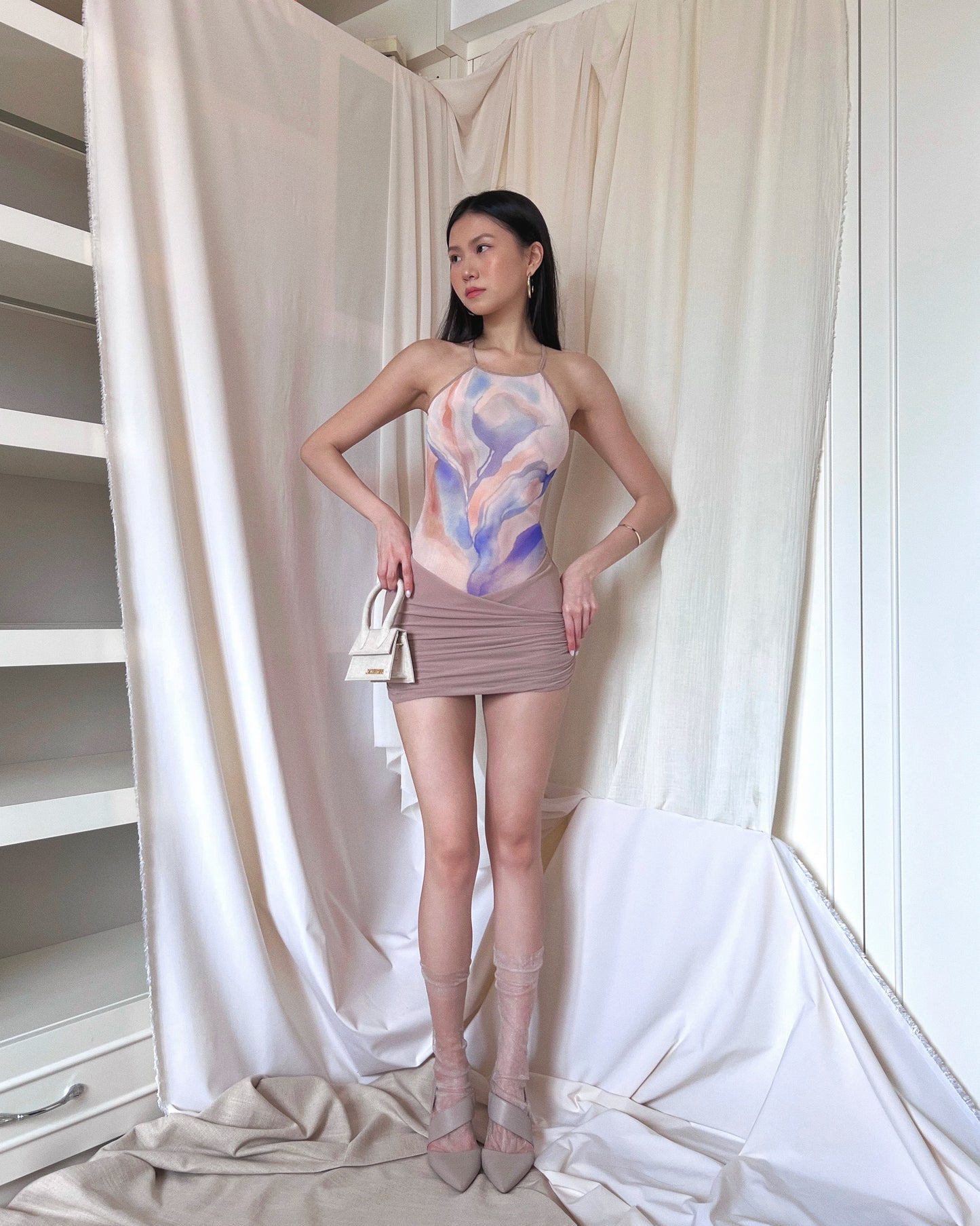 Neutral taupe sleeveless mini dress with floral printed mesh upper body panel with thin straps crossed at back and draped lower part. Styled with Jacquemus mini purse, sheer socks and Calvin Klein heels.