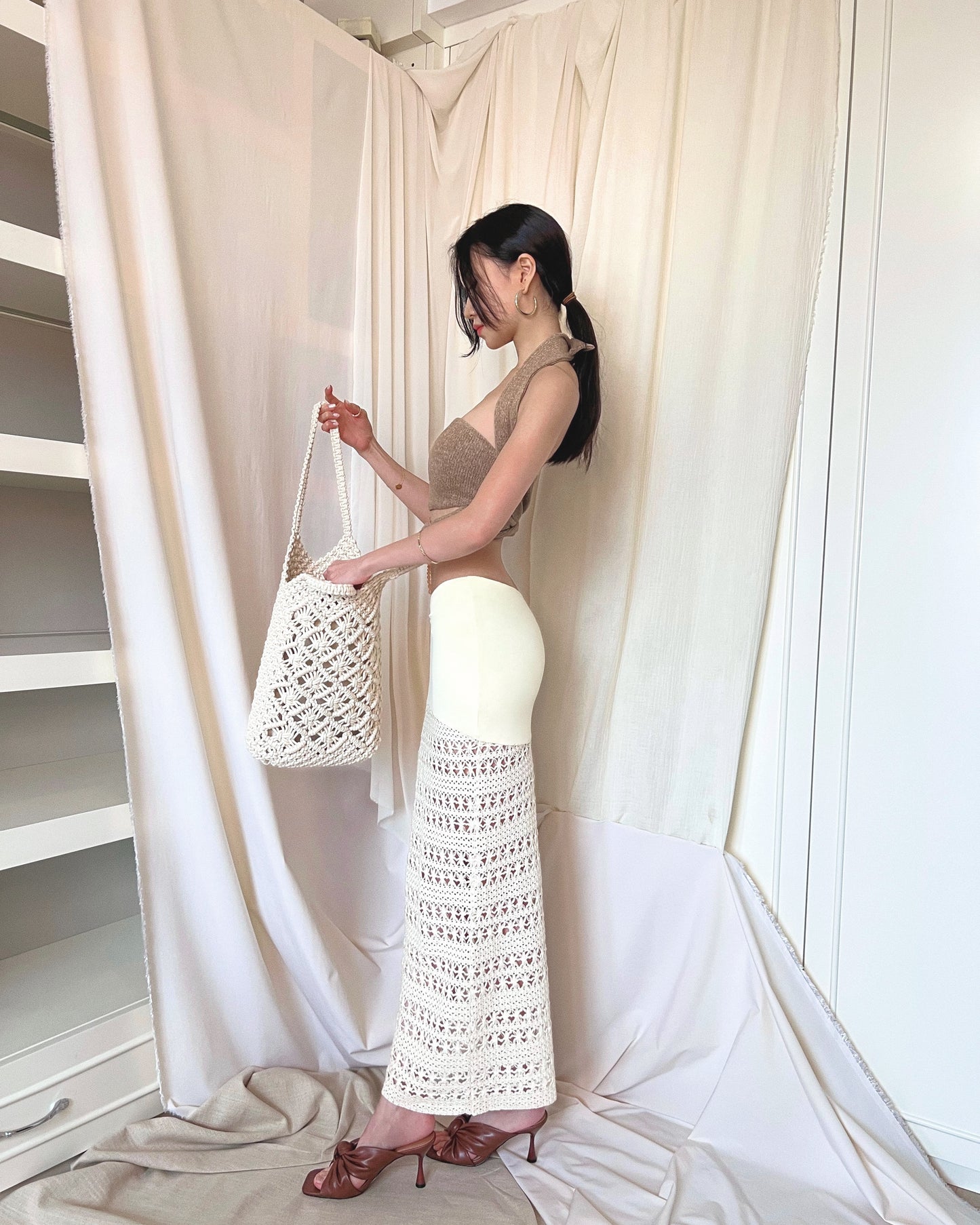 An ivory crochet maxi skirt with nylon fabric and crochet fabric and a symmetrically diagonal waistline. Styled with strapless brown knit top with front twisted knot, long back straps and semi-iridescent beads around waist, and brown mules.